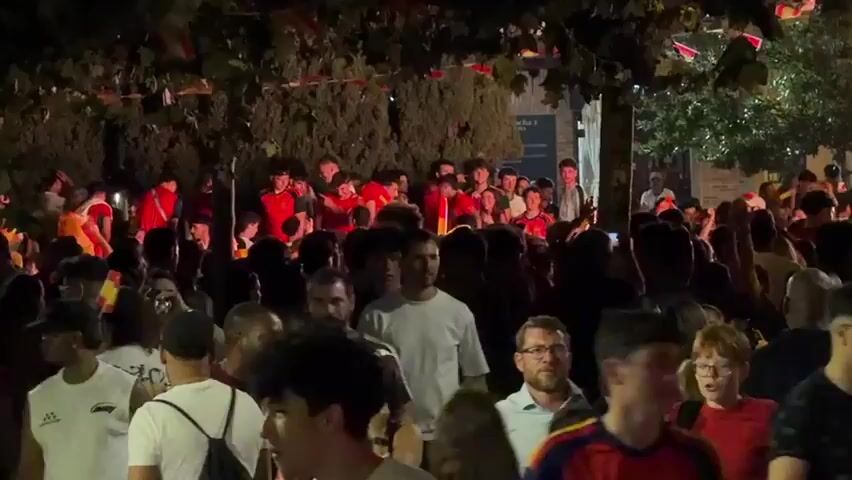 A nighttime outdoor scene depicts a large gathering of people. Many individuals are wearing red shirts. Small flags, predominantly red and yellow, are strung across the upper portion of the frame, suggesting a celebration or event. The crowd appears to be in a public space, possibly a street or square, with trees and some buildings visible in the background. People are standing and moving, with some looking towards the camera and others interacting amongst themselves. The lighting is dim, typical of nighttime, with artificial lights illuminating the area. Some individuals are closer to the camera, while others are further back in the crowd.