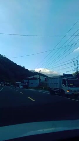 A side view from a moving vehicle captures a road winding alongside a lush green mountain. The sky is a clear blue with a few wisps of white clouds visible above distant mountain peaks. Overhead, a complex network of power lines crisscrosses the frame, supported by utility poles. Along the side of the road, several buildings with blue and gray facades are situated amidst vegetation. Various vehicles, including trucks, vans, and motorcycles, are traveling in the same direction as the camera, contributing to the sense of movement and activity. Yellow road markings are visible on the asphalt, indicating lanes and directions. The overall scene depicts a serene, daytime view of a rural or semi-rural landscape with ongoing traffic.