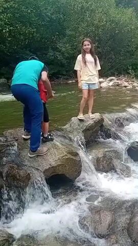 A man in a teal shirt and blue pants is carrying a young child in a red shirt across a rocky stream. The man is holding the child in his arms. To the right of the stream, a young girl in a yellow shirt and shorts stands on a rock. The stream has clear water with visible rocks and white foam from small waterfalls. The background is filled with green trees and foliage. The video is shot from a low angle, looking up at the scene.