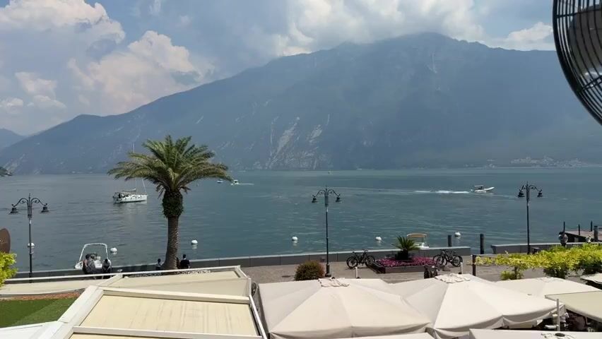 The video opens on a panoramic view of a serene lake surrounded by majestic mountains under a partly cloudy sky. In the foreground, a row of outdoor cafe tables with white umbrellas is visible, some folded and some open. A bright green patch of artificial turf is on the left. A paved walkway runs along the edge of the lake, where a single tall palm tree stands prominently. Several people are seen strolling along the walkway, some seated on benches. A few boats, including a small white one and a larger vessel, are navigating the calm waters of the lake. In the mid-ground, streetlights with ornate designs illuminate the path. Bicycles are parked near a cluster of pink flowers. The background is dominated by imposing, mist-covered mountains that extend as far as the eye can see. The video pans slightly to the left, revealing more of the lake and the mountainous landscape. The overall atmosphere is one of peaceful tranquility and natural beauty.
