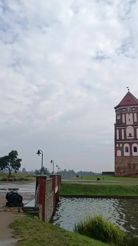The video pans from left to right, showing a view of Mir Castle complex in Belarus. In the foreground, there is a body of water with green reeds growing along the edge. To the left, a brick wall with lampposts leads to a bridge. The castle itself is visible in the background, with its prominent red-tiled roofs and brick walls. The sky is cloudy.