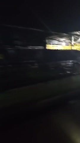 A blurry, dark video pans across an outdoor, open-air market or food stall area at night. Several motorcycles are parked in front of stalls. There are banners with writing and images hanging above the stalls. Some people are visible sitting at tables or standing near the stalls. The ground appears wet, suggesting it may have rained or been recently cleaned. The lighting is dim, with yellow and white lights illuminating the area. The camera movement is fast and jerky, causing significant motion blur.