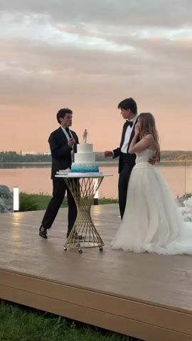 A wedding reception is underway outdoors at dusk. Three people are gathered around a tiered wedding cake on a table. The person on the left, dressed in a black suit and bowtie, is speaking into a microphone. The person in the middle, a young man in a black tuxedo, is holding the microphone and looking towards the person on the right, a young woman in a white wedding dress. The wedding dress is a strapless ballgown with a fitted bodice and a voluminous skirt. The young woman appears to be reacting emotionally, covering her mouth with her hand. The background shows a body of water with trees and a hazy sky with hints of pink and orange, suggesting a sunset. A faint rainbow is visible in the distance. The reception area is set on a wooden deck.