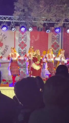 A group of female performers in traditional folk costumes are on a stage at night. The costumes appear to be red and white with intricate patterns. Some of the performers are holding what look like microphones. In the background, there is a red and white patterned backdrop and stage lighting. A crowd of people are visible in the foreground, with their backs to the camera, watching the performance. The overall scene suggests an outdoor cultural event or festival.