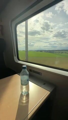 The image provides an interior view from a modern train carriage, looking out a large window at a sunlit rural landscape. In the immediate foreground, a clear plastic water bottle with a blue cap rests on a light-colored, possibly wooden, fold-out table attached to the train seat. The bottle is approximately half-full, and the strong sunlight streaming through the window casts a distinct shadow of the bottle onto the table. Visible text on the bottle's label includes "EV*QUELLE" and "STILL".

Beyond the window, a wide expanse of bright green, undulating fields stretches into the distance under a partly cloudy sky. The landscape suggests a vast agricultural area, possibly moving slightly, indicating the train is in motion. A faint treeline is visible on the horizon, and scattered houses or small buildings can be seen in the far distance, suggesting a small settlement. The sun itself is visible as a bright, almost blinding, light source through the clouds in the upper left portion of the window.

The scene depicts a peaceful journey during daytime, with clear weather. No people are visible, but the presence of the water bottle and table implies a passenger is occupying the seat. The setting, identified as Reichertshausen, Germany, aligns with the European rural scenery observed.