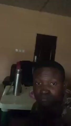 A man is recording a selfie video indoors. He is looking directly at the camera with a serious expression. Behind him, there is a small white table with a silver thermos on it, a black chair, and a dark door in the background. The room has a light orange-colored wall. The lighting is dim.