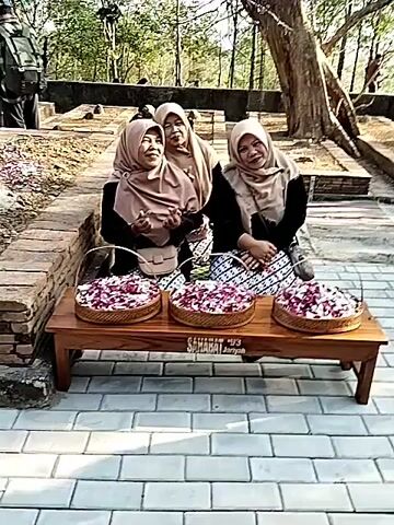Three women wearing headscarves are sitting on a wooden bench. In front of them are three woven baskets filled with red and white flower petals. Behind the women, a tiered fruit display is visible, topped with small flags. Several people in traditional yellow and green attire are seated in rows to the left of the fruit display. In the background, trees and more people are present. The scene appears to be outdoors, with paved ground and brick structures.
