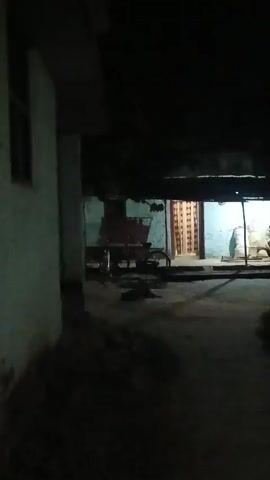This is a night-time, low-light video filmed from a slightly elevated, stationary perspective. The scene appears to be an outdoor courtyard or pathway leading to a building with a porch. The building has a light-colored facade and a dark roof. There is a doorway with a decorative door and an open porch area. Objects visible on the porch include what appears to be sacks or bundles, and possibly some furniture. To the left of the building, a cart-like object with wheels is partially visible. In the background, beyond the building, trees are visible, and a street light illuminates the area. The foreground is dark, with some indistinct grassy or earthy textures. The overall impression is of a rural or semi-rural setting at night, with artificial lighting providing minimal visibility.
