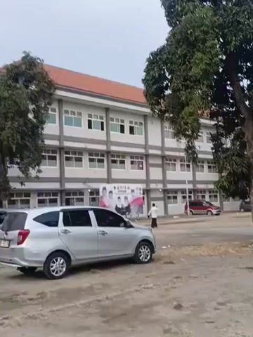 The video shows a silver minivan parked in a dirt lot in front of a large, multi-story building. The building is white with gray accents and has many windows. There is a large banner on the side of the building with images of people. A large tree with dark green foliage is in front of the building, partially obscuring the view of the middle section. Another tree is to the right of the building. Several other cars are parked in the lot, including a red sedan and a blue minivan. The sky is overcast.