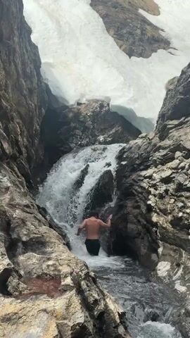 A shirtless man in shorts stands in the shallow water at the base of a waterfall, which is located in a rocky canyon with snow at the top. The man stands facing the camera with arms out at the side, then he puts his face under the waterfall.