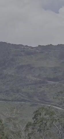 A hazy, wide shot captures a lush, green mountainside with a winding road. At the very top of the mountain, a cluster of buildings is visible, possibly a small settlement or complex. One of the buildings has a flag flying, appearing red and white, suggesting the Indonesian flag. The overall atmosphere is natural and somewhat remote, with a feeling of elevation and distance.