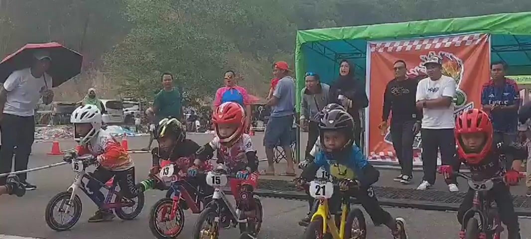 The video shows a children's bicycle race. Several young children in protective gear, including helmets, are lined up on bicycles at the starting line. Some adults are visible in the background, observing the race, with one person holding an umbrella. The race appears to be taking place outdoors on an asphalt track marked with cones and ropes. The children are positioned behind the starting line, indicating the race is about to begin or has just started. The overall atmosphere appears to be one of anticipation and excitement for the young participants.