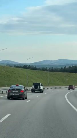 The video shows a view from a moving vehicle, likely a car, driving on a multi-lane highway.  Three cars are visible in the lane ahead: a dark-colored taxi, a white SUV, and a red car further ahead. The highway curves gently. The shoulder of the road has a metal guardrail. Beyond the shoulder is a grassy slope, and further back are hills and some trees under a sky with scattered clouds. Tall streetlights line the side of the road. As the video progresses, the grassy hill changes to show a rocky, disturbed hillside.