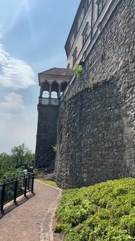 A path curves along the base of a massive stone wall, part of a large building that appears to be a castle. The wall is constructed of irregular, rough-hewn stones of varying sizes and colors, predominantly grays and browns. To the left of the path, a dark metal railing follows its curve, separated from the path by a strip of dirt. Beyond the railing, a lush green hillside slopes downwards, dotted with trees. The path itself is composed of reddish-brown gravel. Above the wall and to the left, a section of the building is visible, featuring windows and a small covered balcony with arched openings supported by columns. This balcony juts out from a tower-like structure built into the side of the main wall. The sky above is a bright blue with wispy white clouds, and the sun casts bright rays and shadows, indicating a sunny day. Green bushes and low-lying plants grow along the right side of the path, directly against the stone wall. The overall impression is one of a historic site, possibly a tourist attraction, with a scenic view.