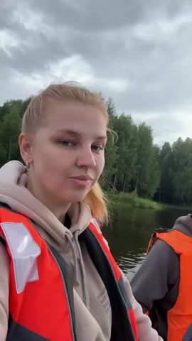A young woman with blonde hair pulled back in a ponytail and wearing an orange life vest in the foreground turns to look at the camera and smiles. Behind her, a man wearing a dark baseball cap and an orange life vest sits in a boat, looking forward. The boat appears to be moving through a body of water, as indicated by the wake behind it. The background shows a treeline of green trees under a cloudy sky.