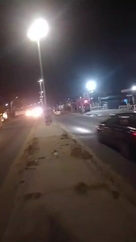 The video shows a view of a road at night. Cars are driving on the road. There are streetlights illuminating the scene. The camera appears to be handheld, and is positioned on a sidewalk next to the road. The sidewalk is dirty. There are buildings and establishments visible on the other side of the road. The video is short and does not show any specific actions or interactions. The environment is urban, with a focus on the road and its surroundings at night.