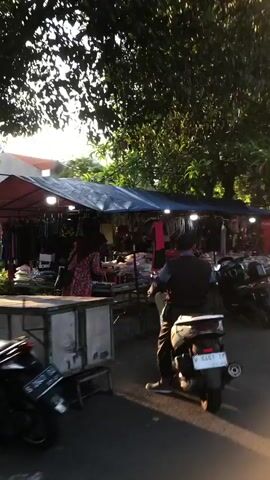 This video captures a scene at an outdoor market in Sumokali, Indonesia. The market appears to be set up along a street, with stalls displaying various goods. In the foreground, a person is riding a scooter, suggesting movement and a dynamic environment. The stall itself is covered by a blue tarp, providing shade for the merchandise and vendors.

The merchandise on display includes what looks like clothing and accessories, with racks of items visible. There are a couple of individuals seen at the market: one person is seated near the stall, possibly a vendor or shopper, and another person appears to be standing and attending to the goods.

The time of day seems to be late afternoon or early evening, as evidenced by the warm, slanting sunlight filtering through the lush green trees that overhang the market area. The presence of trees suggests a tropical or subtropical climate. The weather appears to be pleasant, with no signs of rain.

The overall atmosphere of the market is one of everyday commerce. The scene is bustling and active, typical of a local marketplace where people gather to buy and sell goods. There are no explicit emotional cues that stand out, but the general impression is one of a vibrant community gathering.

Notable details include the scooter in the foreground, the tarp-covered stall, and the abundance of greenery. The sunlight creates dappled patterns on the ground, adding to the visual texture of the scene. The setting is clearly an open-air market in a somewhat informal setting, likely a street market rather than a formal enclosed market.

There is some visible text on a red structure at the right side of the stall, which reads "SCREEN." This could be a brand name or a descriptor of the products sold. The rest of the text on the merchandise or signage is not legible due to the video's resolution and the distance.