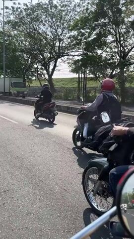 A person rides a motorcycle on a paved road. The rider is wearing a dark jacket and a helmet. Another person is riding on the back of the motorcycle, also wearing a helmet. The motorcycle is black and silver with a license plate that reads "H 8530 TWN". In the background, there is a green truck, trees, and a body of water. The sun is shining, casting long shadows on the road, suggesting it is late afternoon. The scene appears to be in Siring, East Java, as indicated by the location context. The overall mood is neutral, depicting a common mode of transportation in the area.