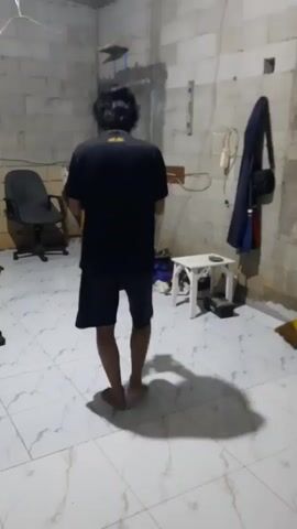 A person is walking away from the camera in a room. The person is wearing a black t-shirt and black shorts. The room has a tiled floor, cinder block walls, and a desk chair. There is a small white table with objects on it and a bag hanging on the wall. The lighting is dim.