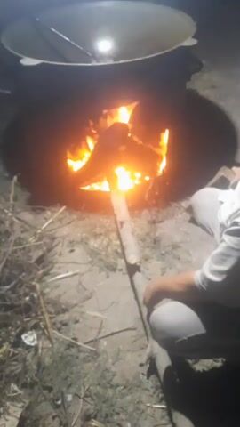 A person is kneeling on the ground next to a large fire pit. The fire is burning intensely with visible flames and embers. A large pot or cauldron is suspended above the fire. The person is holding a long wooden stick and appears to be tending to the fire or adjusting something within the fire pit. The environment appears to be outdoors, with dry ground and some debris scattered around. The lighting suggests it may be nighttime or dusk.