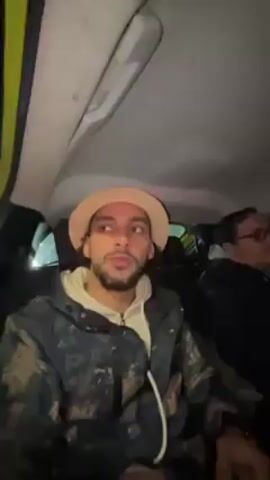 The video shows a group of men in a car. The person filming, who is in the back seat, is wearing a beige hat and a dark jacket with a white hooded sweatshirt underneath. He waves. The driver, who is wearing a black cap and dark clothing, appears to be looking at his phone. The passenger in the front seat is wearing a camouflage jacket. The car is on a street at night, and the dashboard and navigation screen are visible. The overall atmosphere seems jovial.