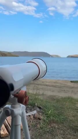 The video shows a close-up perspective of a white spotting scope mounted on a white tripod. The spotting scope is pointed towards a body of water with hills or mountains in the background under a blue sky with scattered clouds. In the foreground, the ground is covered with dry grass and some cut logs. The camera is focused on the eyepiece of the spotting scope, which shows a magnified view of the distant landscape, including the water and a rocky shoreline. The video shifts focus slightly throughout, providing glimpses of both the exterior of the spotting scope and the view through it.