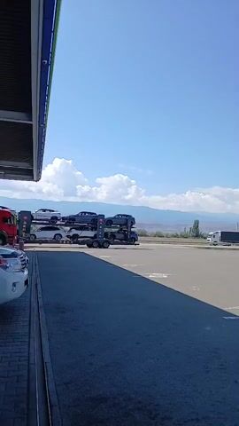 The video pans across a gas station canopy. A white SUV is parked in the foreground under the canopy. Behind the SUV, a car carrier truck is visible, loaded with several cars. Further in the background, a large semi-truck is parked, and mountains are visible in the distance under a clear blue sky with some clouds. The ground is paved asphalt.