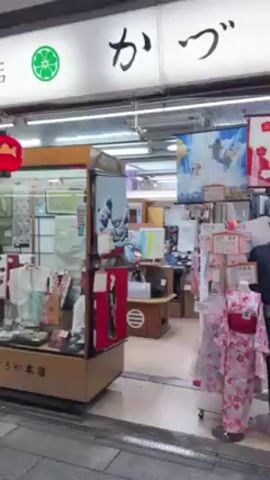 The video shows a street scene in Japan at night.  The camera starts in front of a shop with traditional Japanese decorations, including a green flower symbol and Japanese characters on the sign.  The shop displays various items such as kimonos and other cultural goods. The camera then moves to the street, showing a bustling walkway lined with shops.  People are walking in both directions, some wearing masks. The area is decorated with illuminated cherry blossom-like structures overhead, adding to the festive atmosphere. The overall impression is a lively market scene in Japan at night.