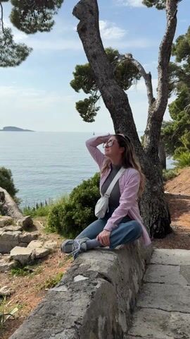 A young woman is seated cross-legged on a stone wall near the coast. She's wearing blue jeans, a gray t-shirt, a pink striped button-up shirt, and gray sneakers with socks. She has sunglasses on her face and is holding one hand up to block the sun. She forms a heart with her hands, smiling as she looks up toward the sky. A white crossbody bag hangs across her body. Behind her, a gnarled pine tree provides shade, and the blue sea stretches to the horizon, where a small island is visible. The weather is sunny and clear.