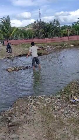 A person wearing a blue hat and jeans bends down by the rocky bank of a shallow river. Another person in a light shirt and dark shorts walks barefoot through the river, holding a stick in each hand. A motorcycle is parked on the far bank, and distant hills and trees are visible under a cloudy sky.