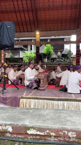 A group of men dressed in white tops and patterned sarongs are playing percussion instruments on a stage. The stage is covered in a red and purple cloth, with a fringed white skirt around the edge. The musicians sit cross-legged, focused on their instruments. They wear purple headbands. Behind them is a covered structure with a tiled roof, open on the sides, revealing buildings and greenery in the background. A large black speaker sits on a stand to the left of the stage. The video is taken from slightly below stage level.