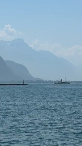 This is a vertical video clip of a serene lake scene with a steamboat sailing across the water. The lake is a deep blue, with gentle ripples on its surface. In the background, a series of layered mountains rise majestically, their peaks softened by the distance and appearing in various shades of blue and grey. A narrow pier with a small lighthouse extends from the left side of the frame into the lake. The sky is a pale blue, with a few wispy white clouds scattered across it.

The steamboat, a classic white vessel with a dark funnel, is moving from left to right across the middle of the frame, leaving a subtle wake behind it. The overall atmosphere is peaceful and tranquil, suggesting a clear, bright day, likely mid-morning or afternoon given the lighting and the presence of the sun. The landscape, with its prominent mountains and lake, strongly suggests a location in the Swiss Alps, aligning with the provided context of Vevey, Switzerland, which is situated on the shores of Lake Geneva. There are no visible people or text in the clip.