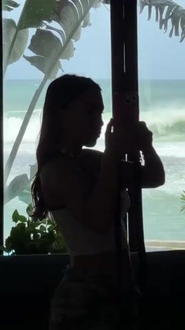 This is a vertical, medium shot video of a young woman taking a selfie in front of a window with an ocean view. She is silhouetted against the bright background, making her features difficult to discern clearly. She has long, dark hair and is wearing a white crop top and camouflage pants. Around her neck is a shell necklace. She is holding a pink smartphone in her right hand, pointed towards a mirror that reflects her image.

The setting appears to be indoors, near a window that looks out onto a beach with waves. Lush green foliage, possibly palm leaves, frame the left side of the window. The sky is a bright blue, suggesting it's daytime, likely afternoon due to the light. The ocean is a vibrant turquoise and white, with visible waves breaking on the shore.

The woman's posture suggests she is posing for the selfie. Her head is tilted slightly upwards, and she appears focused on her phone screen. The overall mood is relaxed and casual, consistent with a beachside vacation or lifestyle. The tile work on the right side of the frame provides an interesting textural contrast to the natural elements. There is no visible text in the video.