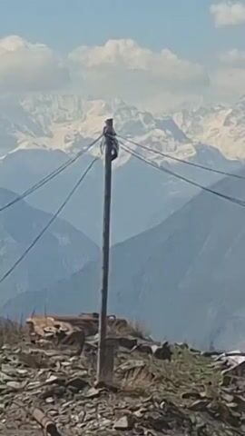 The video shows a landscape of snow-capped mountains viewed from a moving vehicle. In the foreground, there's a wooden utility pole with wires running across the frame. Beneath the pole, there’s a pile of rocks and debris. The road is visible and appears to be winding through the mountainous terrain. The sky is blue with scattered white clouds. As the video continues, the view focuses more on the mountains, showing the snowy peaks in greater detail. The utility pole and wires are visible in the background. The overall impression is of a scenic drive through a mountainous region.