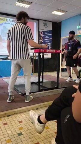 The video shows a man in a black and white striped referee shirt bending down to adjust the table for an arm wrestling match. Another referee in a similar shirt stands beside him. A bearded man in a blue, black, and red shirt stands near the table, waiting to compete. Behind the arm wrestling table, a screen displays text in French. Audience members are visible in the background and in the foreground, observing the preparation. The event appears to be taking place indoors. There are various weights and exercise equipment in the periphery. The floor is covered in a multi-colored checkered tile.