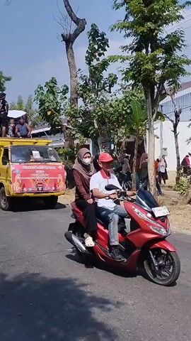 The video shows a street scene with several people and vehicles. In the foreground, on the right, a person wearing a hijab and a brown top is riding a red motorcycle. To the left of the motorcycle, a yellow and red van is visible with people sitting on top. Several trees line the street, and there are buildings in the background. Many people are standing on the side of the road, some are talking, and some are looking at the van. A large red and white flag is planted on the side of the road. The weather appears to be sunny.