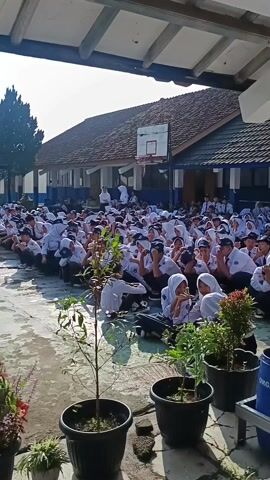 Here is a description of the video:
The video shows an outdoor scene with a large group of students, dressed in white shirts and dark pants/skirts, sitting on the ground in what appears to be a school courtyard. Some of the students are wearing headscarves and/or hats. The students are facing forward and are interacting with one another, some appear to be laughing and chatting.
The courtyard is surrounded by school buildings, with a basketball hoop visible in the background. There are also potted plants and a blue "ORGANIK" barrel visible in the foreground. The camera moves to show more students, who are in line, being inspected by adults in brown shirts.