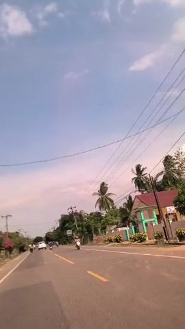 The video shows a road under a blue sky with scattered clouds. The road has a yellow line down the center.  There are cars and scooters driving on the road. Along the road are power lines, trees, and some buildings. There are palm trees visible. A bush with pink flowers is seen at one point.