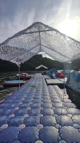 A video shows a floating dock made of blue modular plastic tiles extending into a body of water. The dock is covered by a white mesh canopy that is supported by poles. On the left side of the dock, there are red and blue inflatable water toys. On the right side of the dock, there are several inflatable water structures, including slides and other obstacles. The water is calm, and the sky is cloudy with the sun visible. In the background, there are green hills covered in trees. The camera is mounted on a drone or camera rig that is moving forward, looking down the dock.