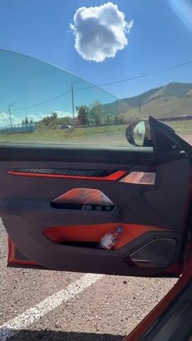 The video begins with an exterior view of a car's open driver's side door, showing the orange and grey interior with a water bottle in the door pocket. The camera then pans inside the car, focusing on the steering wheel and the dashboard screens. The screen displays information including speed, battery status, and a 3D model of the car. The exterior environment is sunny with a blue sky and some hills in the background.