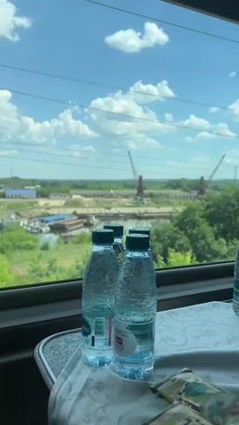 The video pans across the interior of a train compartment, showing bottles of water on a tray on a table. Outside the window, the landscape is visible. There are green trees and a body of water, possibly a river or port. Two large industrial cranes are visible in the distance, along with some industrial buildings. The sky is blue with scattered white clouds. The train appears to be moving, as indicated by the subtle movement and the presence of overhead power lines.