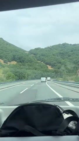 The video shows a first-person perspective from inside a vehicle traveling on a highway. The road is paved with multiple lanes, marked by white lines. On either side of the highway are guardrails. In the distance, two white vans are visible driving in the same direction. The landscape is dominated by lush green hills covered in trees, suggesting a rural or mountainous environment. The sky is overcast with clouds. In the foreground, a black bag is partially visible on the dashboard or passenger seat. The video is characterized by continuous motion as the vehicle drives forward.