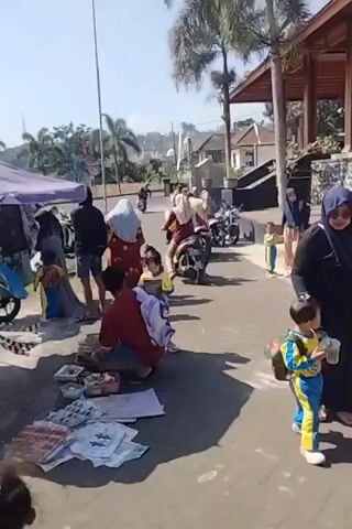 The video shows a street scene with several people and motorcycles. On the left, a vendor is selling items from a stall under a purple canopy. A man in a red shirt and a woman in a blue headscarf are looking at the items. There are two young children, one in a yellow and blue outfit, standing near the vendor. In the background, several motorcycles are parked or being ridden. Further down the street, a woman is riding a motorcycle with a child in front of her. On the right side of the street, there is a building with a wooden roof structure. More people, including children, are visible walking or standing. The ground is paved with stones. Palm trees are visible in the background. The overall atmosphere appears to be that of a marketplace or a busy street on a sunny day.
