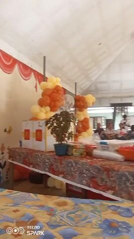 The video shows an indoor gathering, possibly a party, with a group of people. In the foreground, a table is laden with food items and decorations, including two large red plastic containers filled with food, possibly fruits or vegetables, and several boxes with the McDonald's logo. A potted plant is placed in the center of the table. A balloon arch, adorned with orange and yellow balloons, is set up behind the table, anchored by poles. In the background, several individuals are seen interacting, some sitting and others standing. The environment appears to be a covered outdoor space or a large room with a high, sloped ceiling and exposed beams, with fans mounted on the ceiling. The lighting is natural, suggesting daytime.