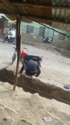 A person crouches near a motorcycle parked on a dirt road. Buildings and other motorcycles are visible in the background. The scene is taken from under a corrugated roof structure.