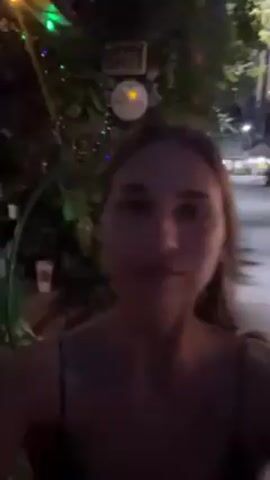 A person with light skin and brown hair is standing on a sidewalk at night. They are wearing a dark top with thin straps and are looking directly at the camera. To the left of the person, there is greenery with colorful lights strung through it. A street with dim streetlights and buildings is visible in the background. A yellow scooter is parked on the left side of the frame. The person looks slightly to their left and back to the camera.