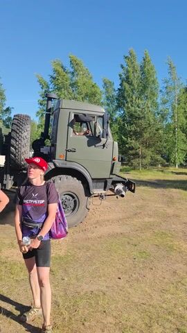 A military truck is being driven in a grassy, wooded area under a clear blue sky, suggesting a daytime event. The truck is olive drab and has large, rugged tires, indicating it is designed for off-road use. It appears to be kicking up dust as it moves. In the foreground, a person wearing a red hat, black t-shirt, and shorts is partially visible, facing away from the camera. They are holding a purple bag. The scene suggests an outdoor activity, possibly a military exercise or demonstration, given the nature of the vehicle and the rural setting. The weather seems to be warm and sunny. The text "6253 AH 51" is visible on the rear of the truck.