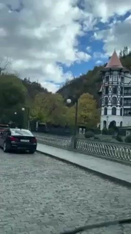 The video shows a view from a car driving on a cobblestone street lined with parked cars. On the left, there's a building with a distinctive turret, followed by a row of shops with red awnings displaying various merchandise. The environment includes trees and a hillside in the background, under a sky with scattered clouds. The camera's point of view moves slowly along the street, capturing the scene.