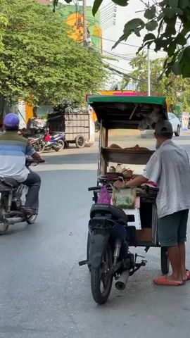 The video captures a daytime street scene in Ho Chi Minh City, Vietnam. In the foreground, a street food vendor stands beside his mobile food cart, which is attached to a Honda motorcycle. The cart features a display case with bread or buns, and various ingredients like fresh vegetables and chili are visible in bags and containers on the counter. The vendor, wearing a hat and a light-colored patterned shirt, gestures slightly while looking towards the street.

Throughout the short clip, motorcycles pass by on the asphalt road. One motorcyclist, wearing a purple helmet, drives past the cart towards the left of the frame. In the background, several other motorcycles are parked along the roadside, and a large trailer or truck loaded with what appears to be garbage is also visible. Lush green trees line the street, and a large building with yellow and orange painted details can be seen further in the distance. The scene is brightly lit by natural sunlight, indicating clear weather.