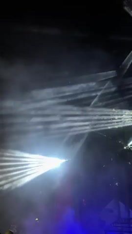 The video captures a nighttime outdoor scene illuminated by numerous bright, white laser light beams. Some beams are sharp and distinct, while others are diffused by smoke or haze, creating ethereal patterns in the dark sky. In the lower portion of the frame, there is a crowd of people visible, their forms silhouetted against the vibrant lights. They appear to be gathered for an event, possibly a concert or festival, as suggested by the stage-like lighting and the presence of a large audience. The overall atmosphere is one of excitement and dynamic visual spectacle, dominated by the interplay of light, smoke, and the shadowy figures of the attendees.